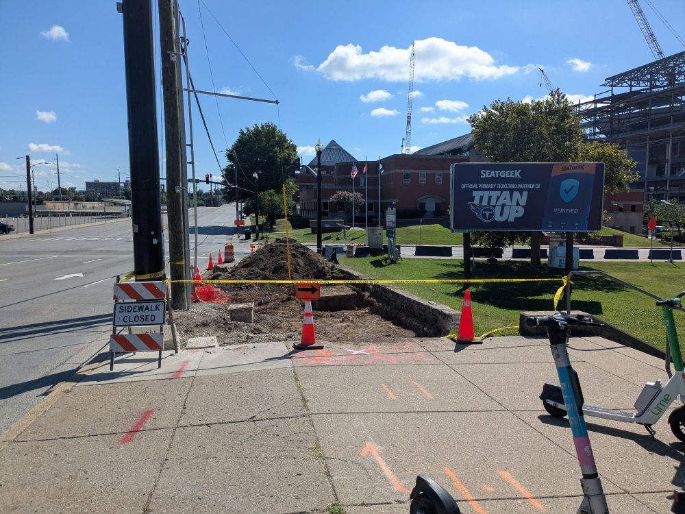 A picture of a closed sidewalk, with a Titans bill board in the background, next to a six lane road, with rental e-scooters in the foreground.