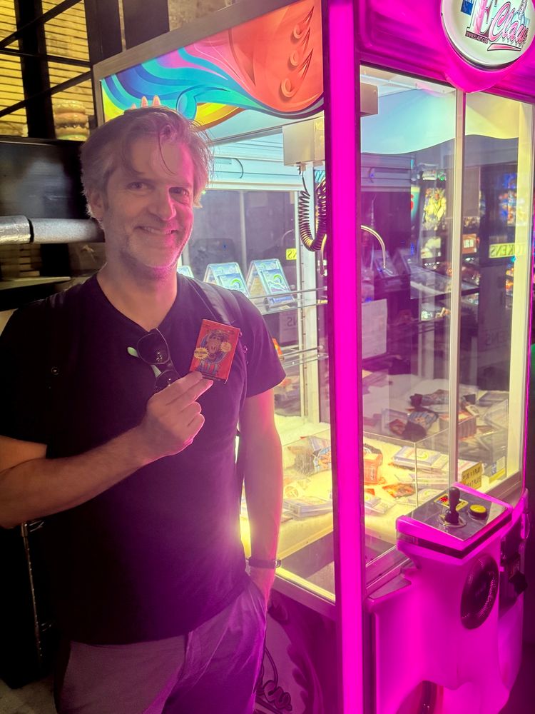 Patron holding up a pack of Alf cards while standing next to a claw machine.