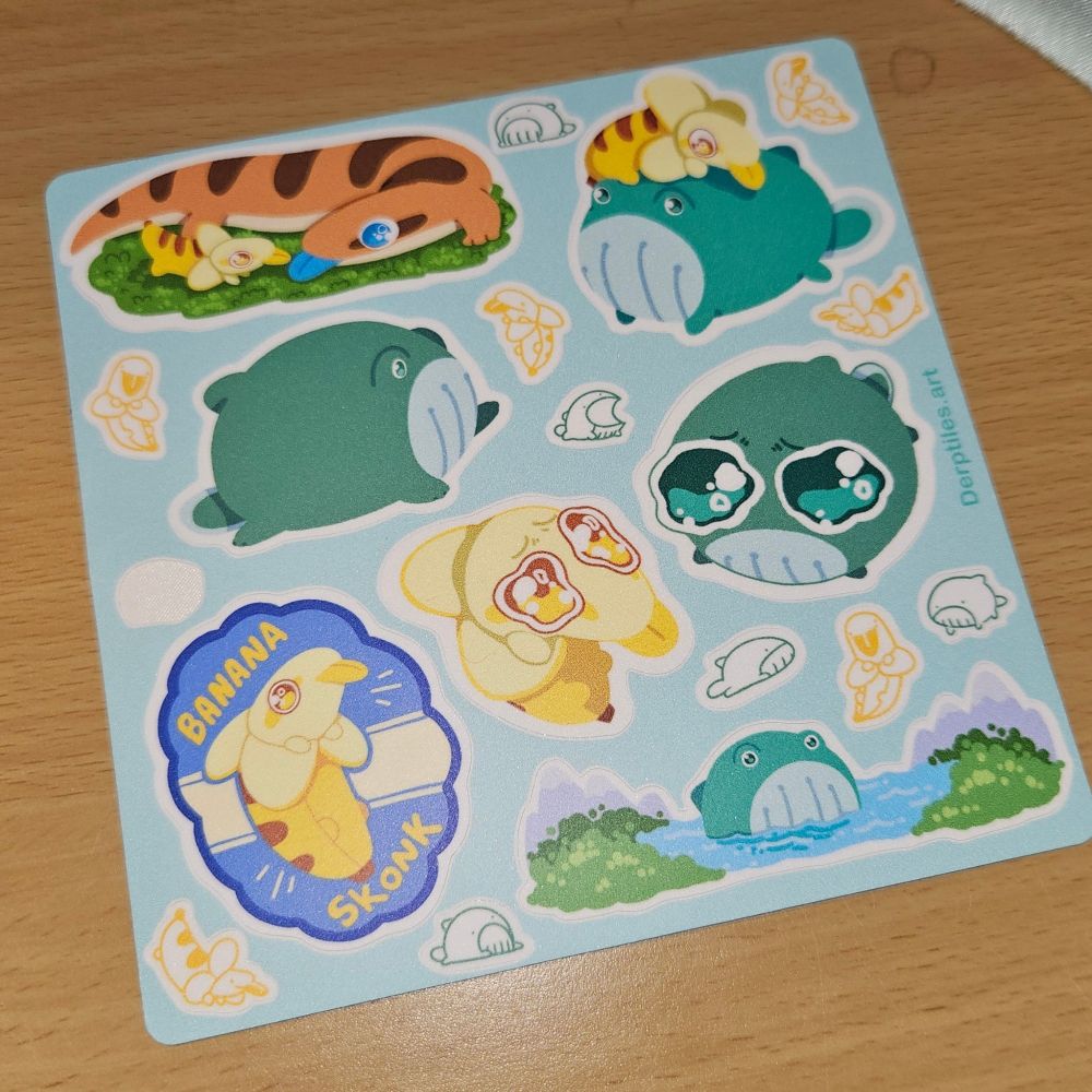 A blue sticker sheet with various illustrations by Derptiles Art depicting their Skonk, Frogwhale, & Banana Skonk characters. One small doodle sticker has already been peeled off on the left side and used. The background is a tan/beige wooden desk.