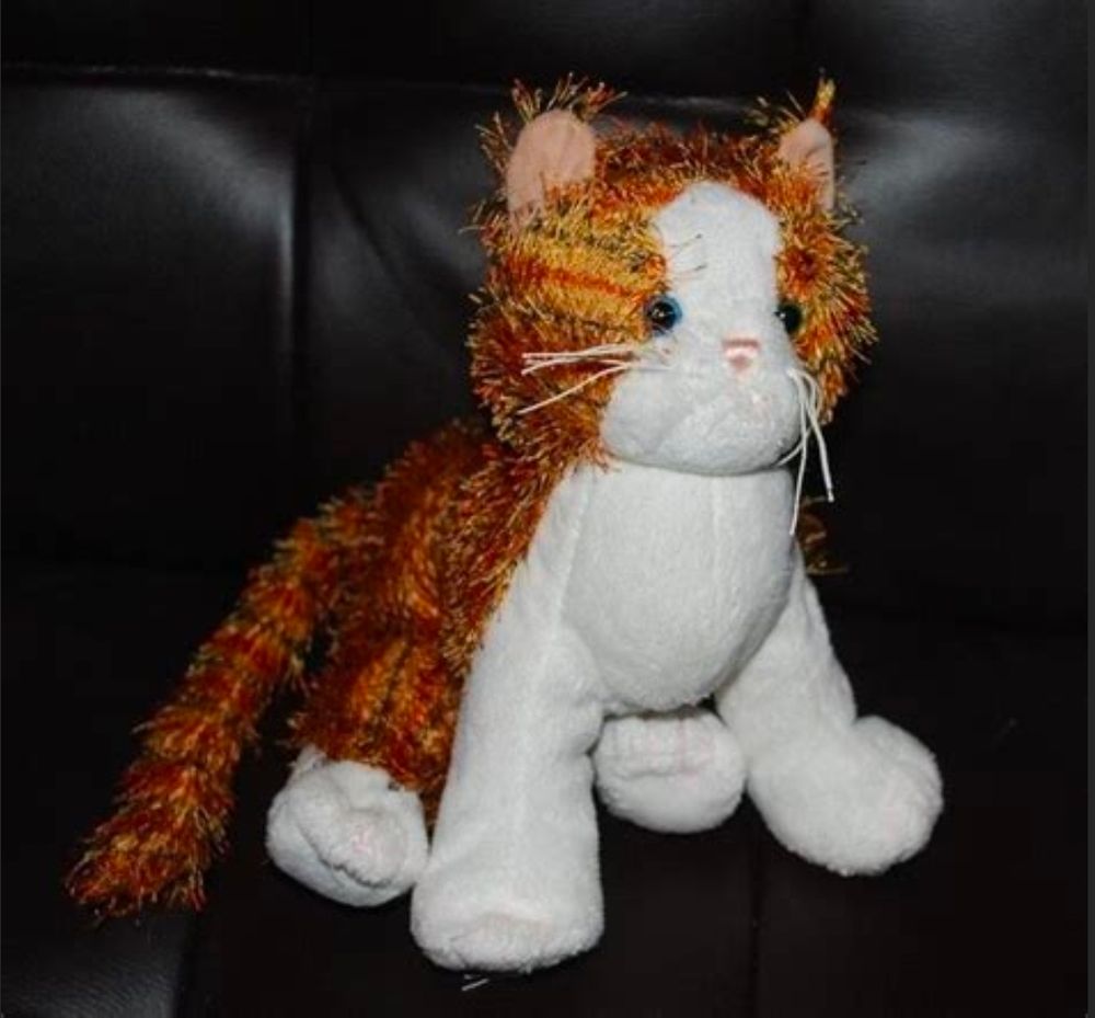 A plushie cat with white paws, face, & underside but with orange & black stripes. It also has two different eye colors - one blue, and one green. It has white string whiskers and pink ears. It is sitting on a black shiny couch.