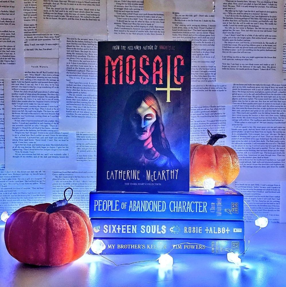 A stack of books with fairy lights and two pumpkins. The books are Mosaic, People of Abandoned Character, My Brothers Keeper and Sixteen Souls.