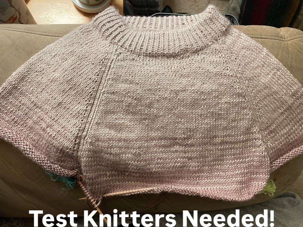 Picture of a sweater in progress. It fades between three colors: cream, lavender, and dark purple. The picture shows the first color (cream) finished and faded into the second color (lavender), displayed on the armrest of a chair. There is text at the bottom of the picture that says “Test Knitters Needed!”
