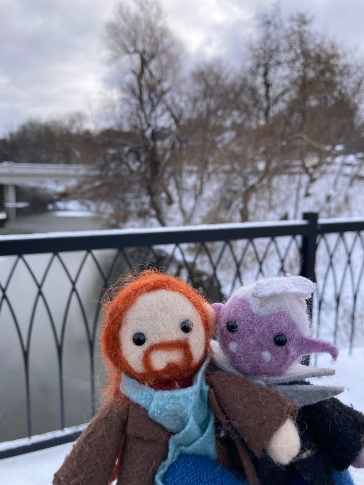 small felted toys of Caleb Widogast and Essek Thelyss are outside on a snowy day. They are on a bridge, with a black iron handrail behind them. Beyond is a view of a cold-looking river and a snow-covered, tree-lined slope. Caleb is wearing a scarf; slightly behind him, Essek looks like he’d rather be indoors.  