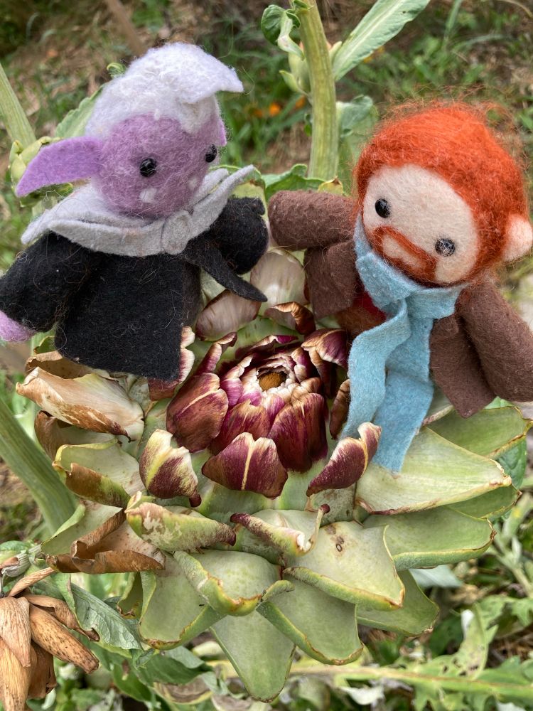 Small felted characters of Essek Thelyss and Caleb Widogast from Critical Role sit on a blooming artichoke’s symmetric petals, looking down at the purple centre of it. 