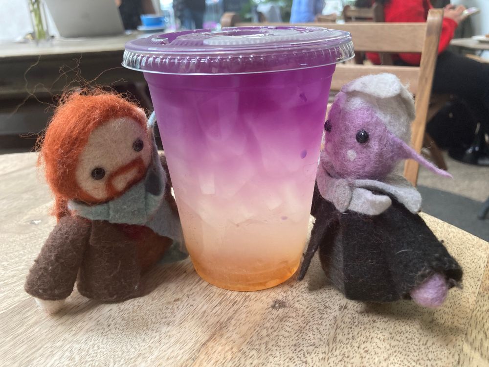 Small felted toys of Caleb Widogast and Essek Thelyss stand on either side of a plastic cup. It is full of an iced drink that starts a vivid purple at the top, then fades through white to orange at the bottom. 