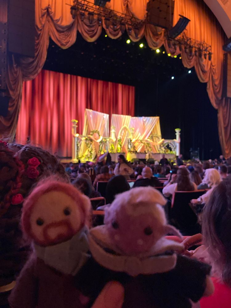 Small felted toys of Essek and Caleb are held up in front of a stage. The set is decked out in pink draperies and flowers and green leaves