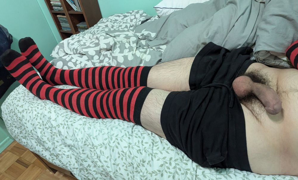 Man laying in bed wearing red and black striped over the knee socks, black shorts, and has an erect penis