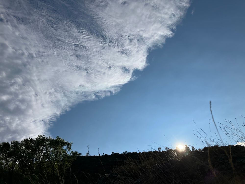 Soleil couchant sur une petite colline dans l’ombre, ciel encore bleu et nuages dans un tiers gauche du cadre, qui font un peu comme des ridules à la surface de l’eau