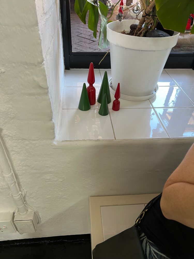 A handful of small, wooden Christmas decorations sit on a stark white windowsill, two red, three green. The green ones are simple cones of different heights. The red ones... are rounded cones on flared bases.