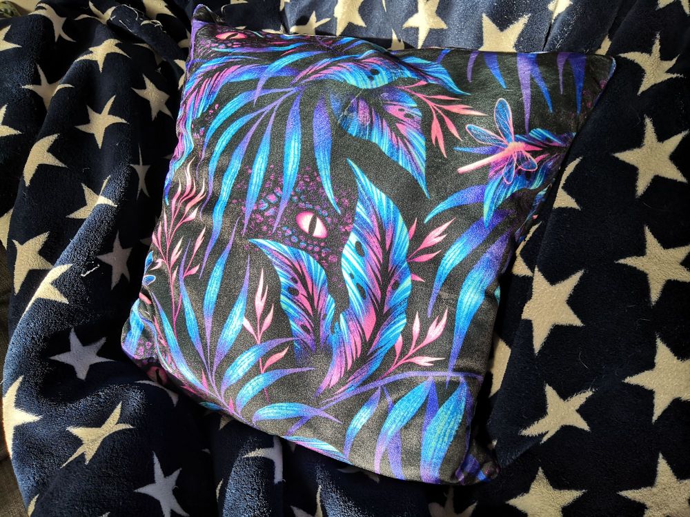 Le superbe coussin "Discreptile", des feuilles type jungle bleues et rose fuchsia avec au milieu un œil de dinosaure qui nous observe, bien caché