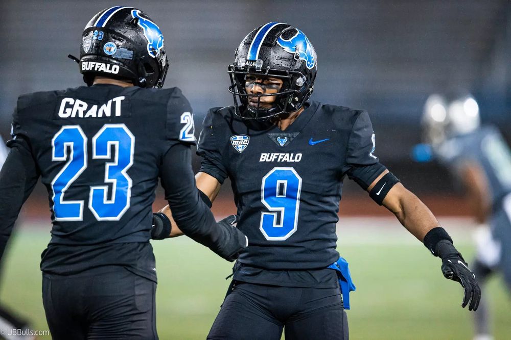 Two University at Buffalo Bulls football players in black uniforms and helmets with blue lettering. 