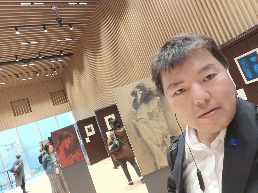 スーツ姿の榊原平氏が「怪談」展の会場内で自撮りをしている。背景は木目調の壁が特徴的な展示空間で、モノクロの人物画や赤い絵画などの作品、他の来場者の姿が見える。