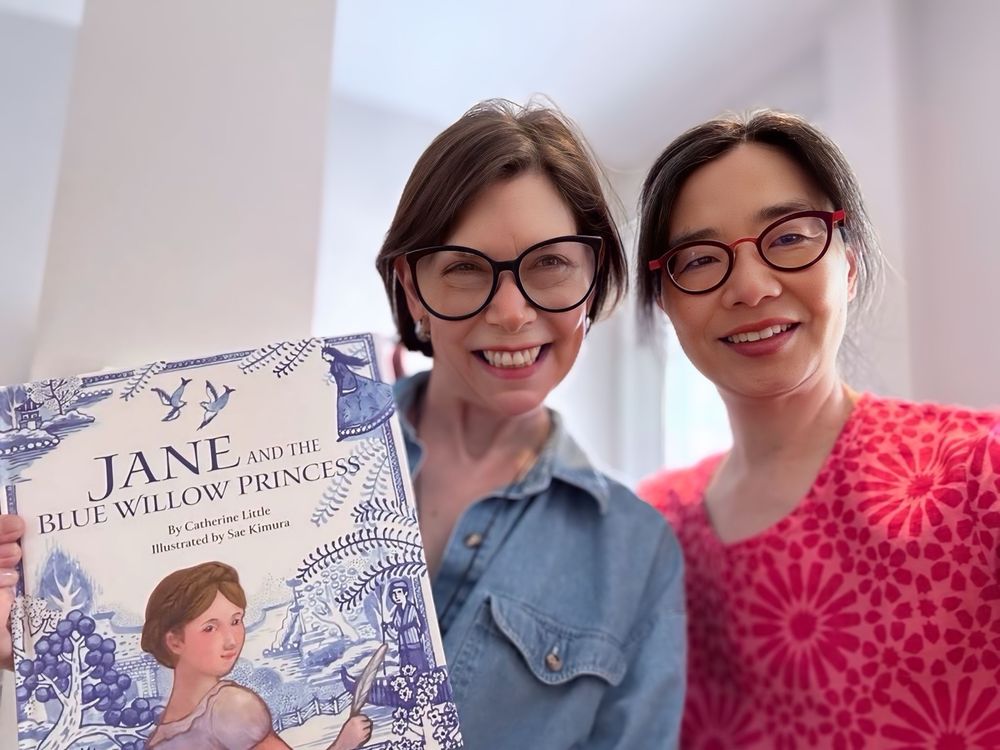 Two smiling women. One is holding up a book titled Jane Austen and the Blue Willow Princess. The book cover portrays a young Jane Austen surrounded by a lovely blue pastoral scene. 