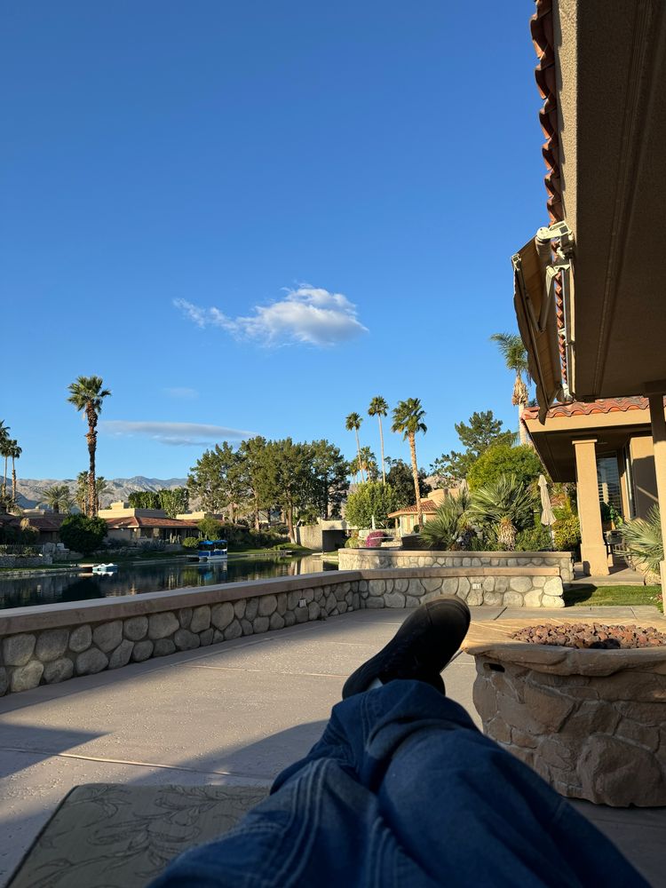 Lounging legs on a patio, under blue skies with palm trees and a manmade lake. 