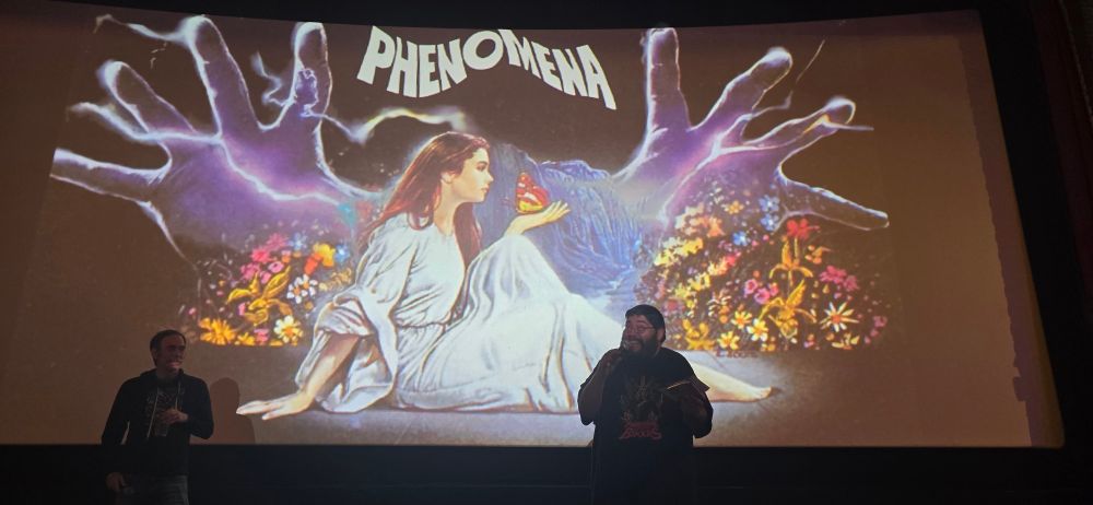 Greg D and Jenius McGee of the Nightmare Junkhead podcast stand in front of a cinema screen with the promotional poster of Phenomena on it.