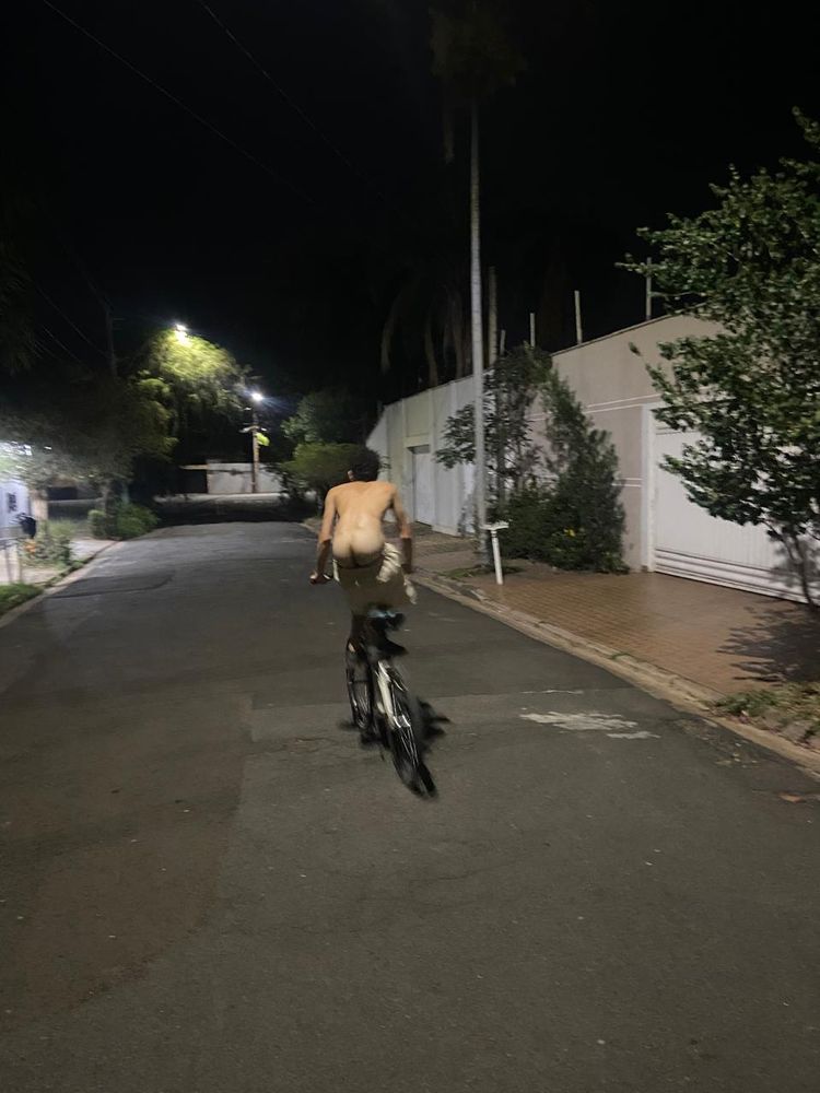 Homem cis em cima de uma bicicleta em movimento. O homem está com a bunda de fora do short e sem camiseta. A rua é larga e está sem carros.