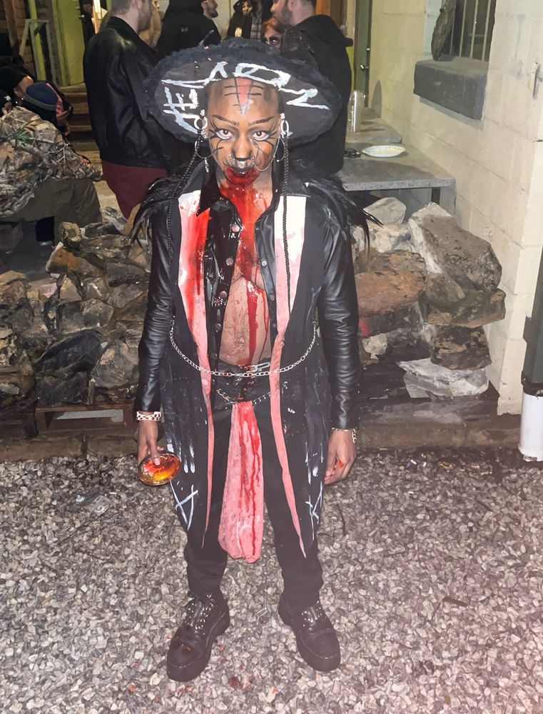 Oberon the Demon freshly covered in blood. They’re wearing an all black ensemble that includes a hat with “DEMON” on the inner rim and a trench coat length feather vest