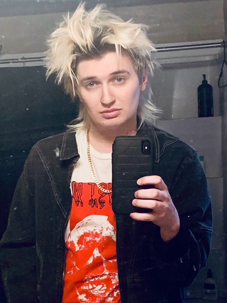 blonde guy with messy hair a fake gold chain and a denim jacket on over a red and white t shirt 