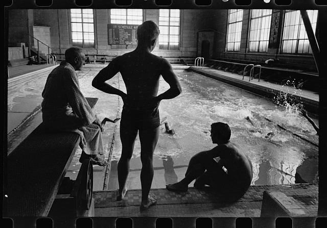 Black and white photograph by Stanley Kubrick for LOOK magazine. An indoor swimming pool with three men in the foreground, silhouetted by the light of the pool. A coach in plainclothes sitting on a diving board, a swimmer student standing with arms on his hips, and another swimmer sitting at the edge of the pool.