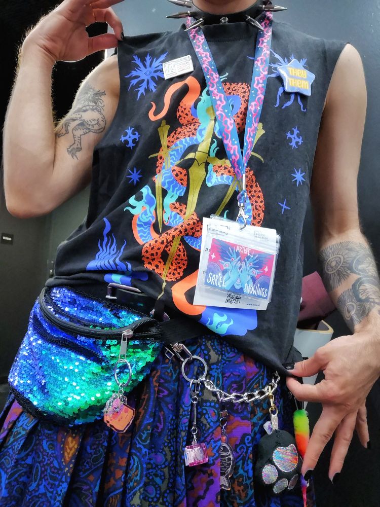 A picture of Sariel's torso showing off their outfit. They're wearing a skipek collar, a sleeveless t-shirt with a drawing of a red and blue neon snake coiled around three golden swords, a colourful patterned skirt in deep blues and purples, a belt and a chain with a variety of charms hanging from it and an iridescent fanny pack covered in sequins. 