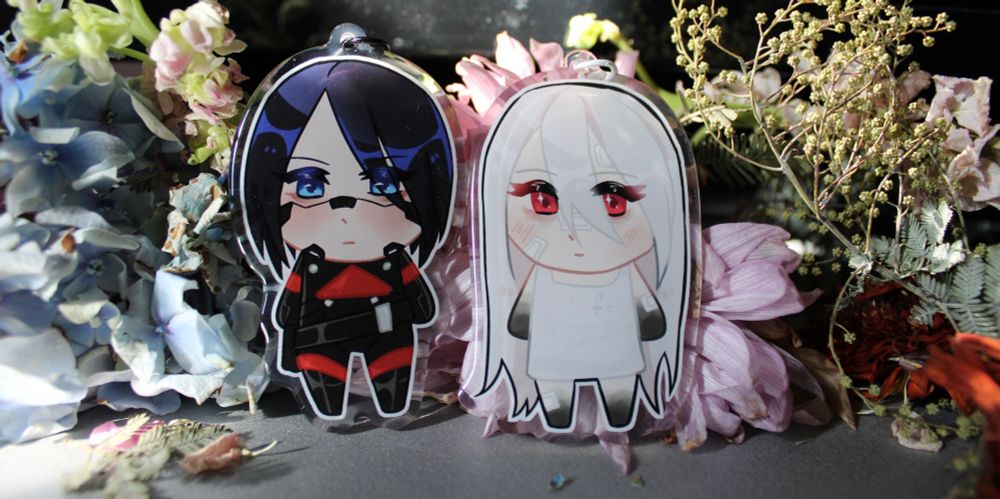 Elster and Ariane keychain next to eachother surrounded by blue, purple and red flowers. 