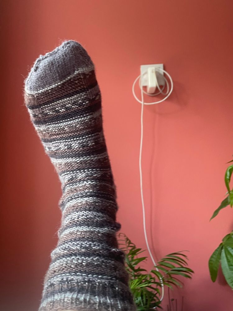 A hand-knitted sock in grey, black and brown against a coral coloured wall. 