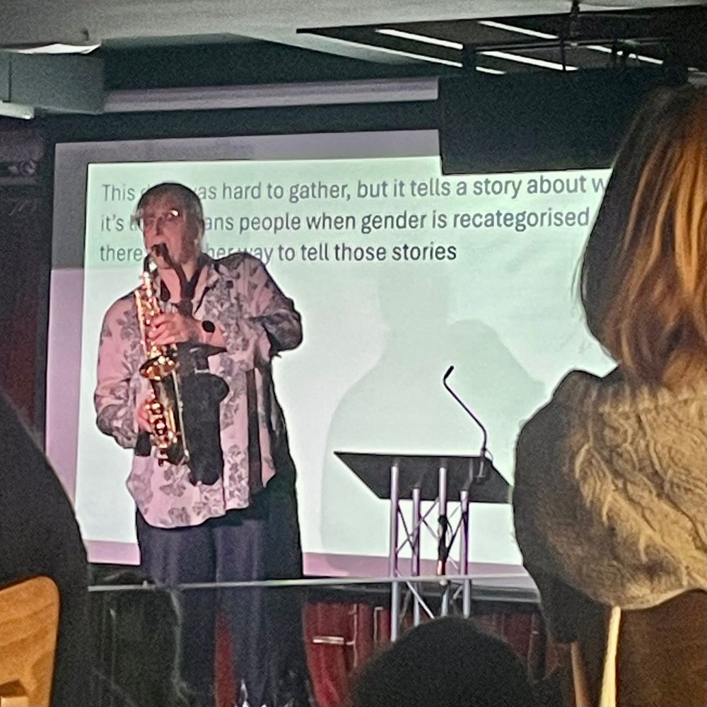 A tall white woman in a floral short playing saxophone in front of a PowerPoint presentation. Black text on a white background reads: The data was hard to gather, but it tells a story about wh… it’s… trans people whose gender is recategorised… there… a way to tell those stories.  
