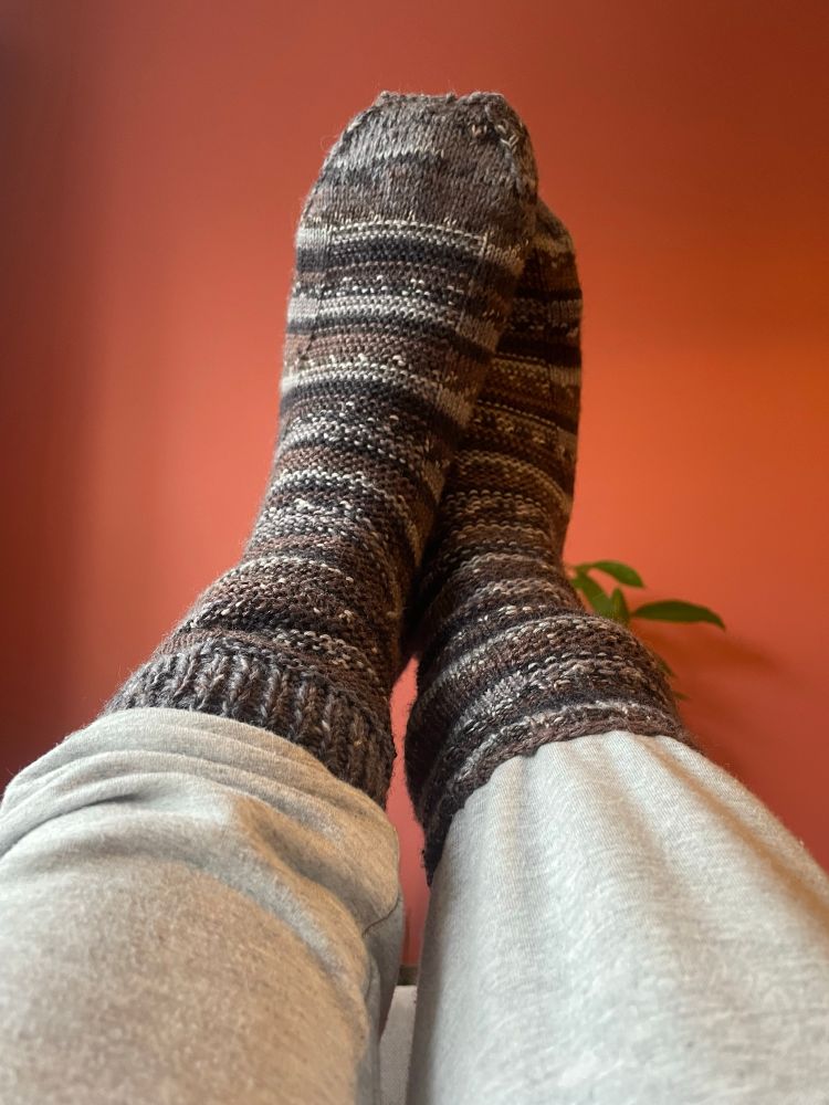 Light grey tracksuits tucked into two hand-knitted socks in grey, black and brown, photographed against a coral coloured wall. 