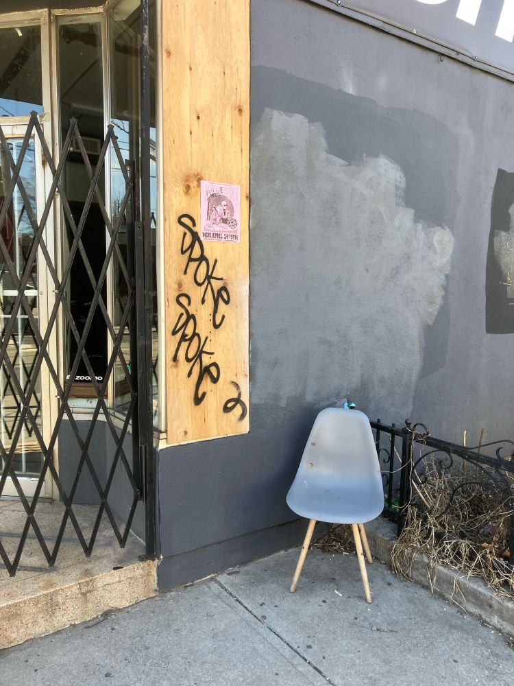 Grey molded chair in wooden legs outside a closed up shop. Plywood with black graffiti “spoke spoke” is written. Sunny spring light. 