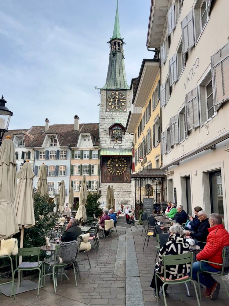 The old clocktower in the center of Solothurn and a half empt terrace with some people having coffee or tea and of course a slice of cake. 
It's a warm and sunny Spring day, but people are still in a bit of a Winter mood with their jackets on.
