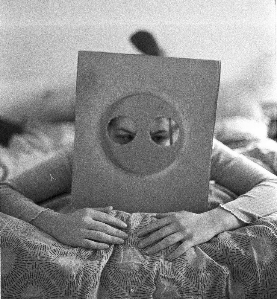 Self-portrait in black and white with square faced mask. Person is laying on the bed 