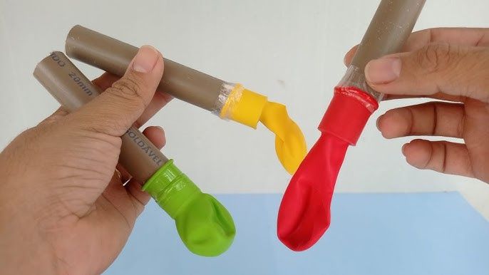 duas mãos segurando três pedaços de cano PVC com balões de festa vazios presos em um de seus lados com fita durex
