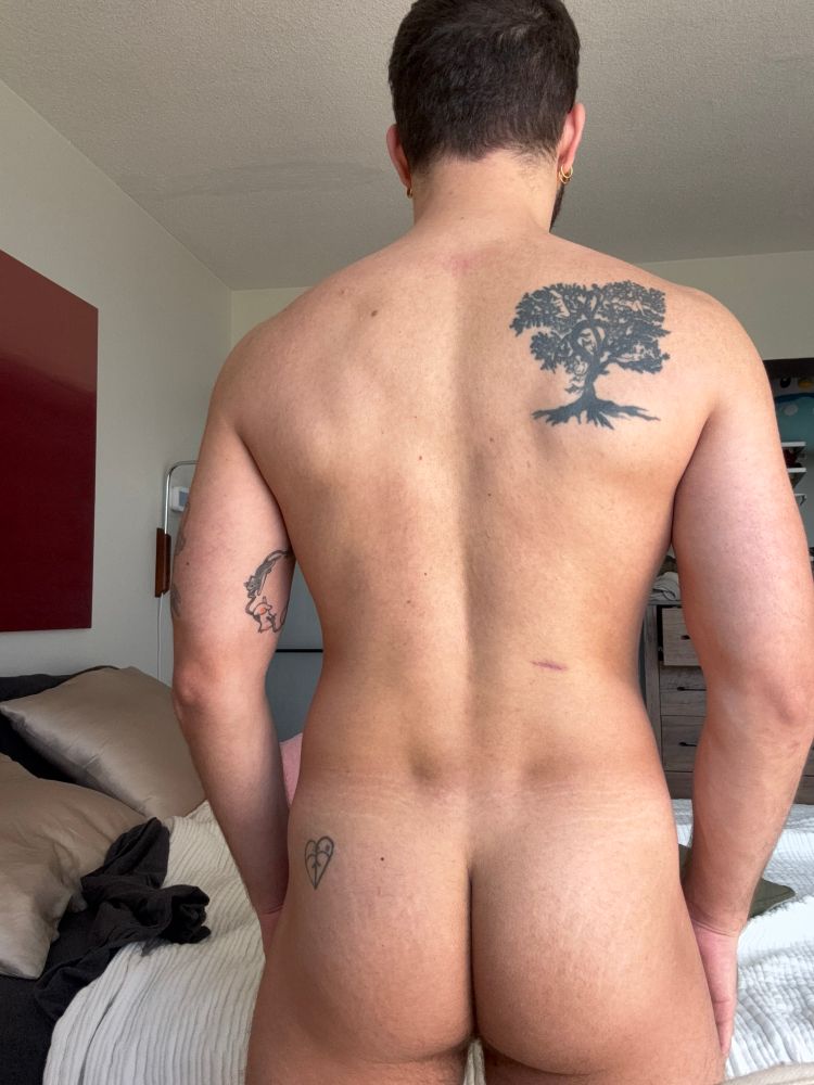 Picture of a gay’s nakey back and butt with a visible heart tattoo on their butt, tree on their back, and cats on their inner bicep 