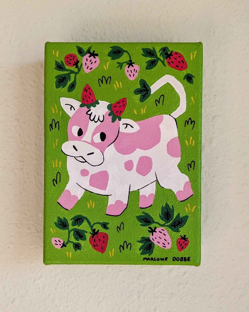 A photograph of a 5x7" canvas, it's painted bright green an has a cartoonish pink cow with strawberries for horns. There are a handful of strawberry plants surrounding it.