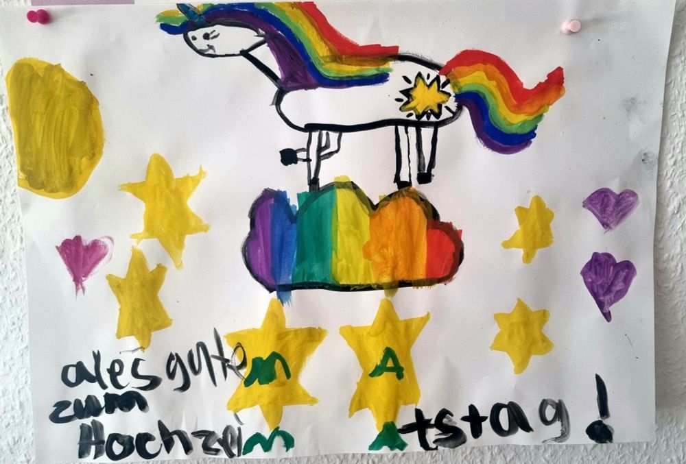 Mit Wasserfarben gemaltes Kinderkunstwerk. Weißer Hintergrund, gelbe Sterne, ein Einhorn mit Tegenbogenmähne, das auf einer Regenbogenwolke steht. Darunter der Schriftzug "Alles gute zum Hochzeitstag Mama"