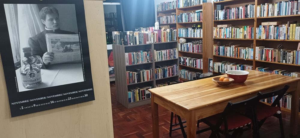 View into a room lined with books (volumes of fiction alphabetised by author surname, and the ones in frame are from Willa Cather through F. Scott Fitzgerald), a table with two fruit bowls, and a Calendario Romano in the foreground open to November, showing a man dressed as a priest reading a copy of El Pais as light streams in through a window behind him to his right.