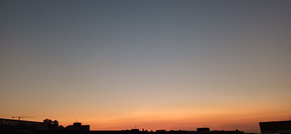 The light of the setting sun refracted through dust and pollution looking southwest along the Witwatersrand, with Charlotte Maxeke public hospital on the left, the broadcast tower in Brixton next to the SABC, South Africa's public broadcaster in the middle of the frame along the bottom, and building rooftops to the right; the sky is cloudless.