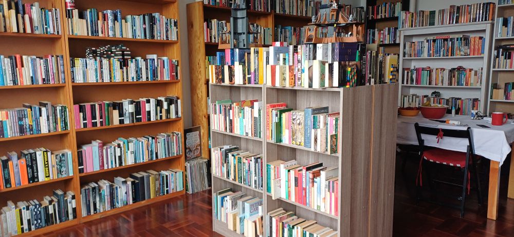 Volumes of fiction on various shelves and bookcases surround a table covered with a white cloth on which two bowls of fruit and a red mug of tea can be seen.