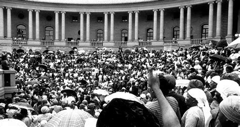 The women's gathering in the amphitheatre of the Union Buildings on 9 August 1956, with government offices visible behind the pillars, and white apartheid civil servants and officials looking on from the walkway behind the balustrades. The Union Buildings, constructed between 1911 and 1913, designed by British colonial architect Herbert Baker (who also designed State House in Nairobi, and several notable government buildings in New Delhi), became the seat of the unified white supremacist colonial government established in 1910 between the four colonialist white supremacist settlements (the Boer republics of the Zuid-Afrikaansche Republiek and the Oranje Vrijstaat; and the British Cape and Natal colonies), in the city named after the first president of the white supremacist republic established by the descendants of Dutch settlers and other European colonists established in 1852. In 1948 it became the seat of the apartheid government, and in 1994 the seat of the post-apartheid democratic government. The amphitheatre is also the space in which Nelson Mandela took the oath of office to become the first president of a democratic Republic of South Africa.