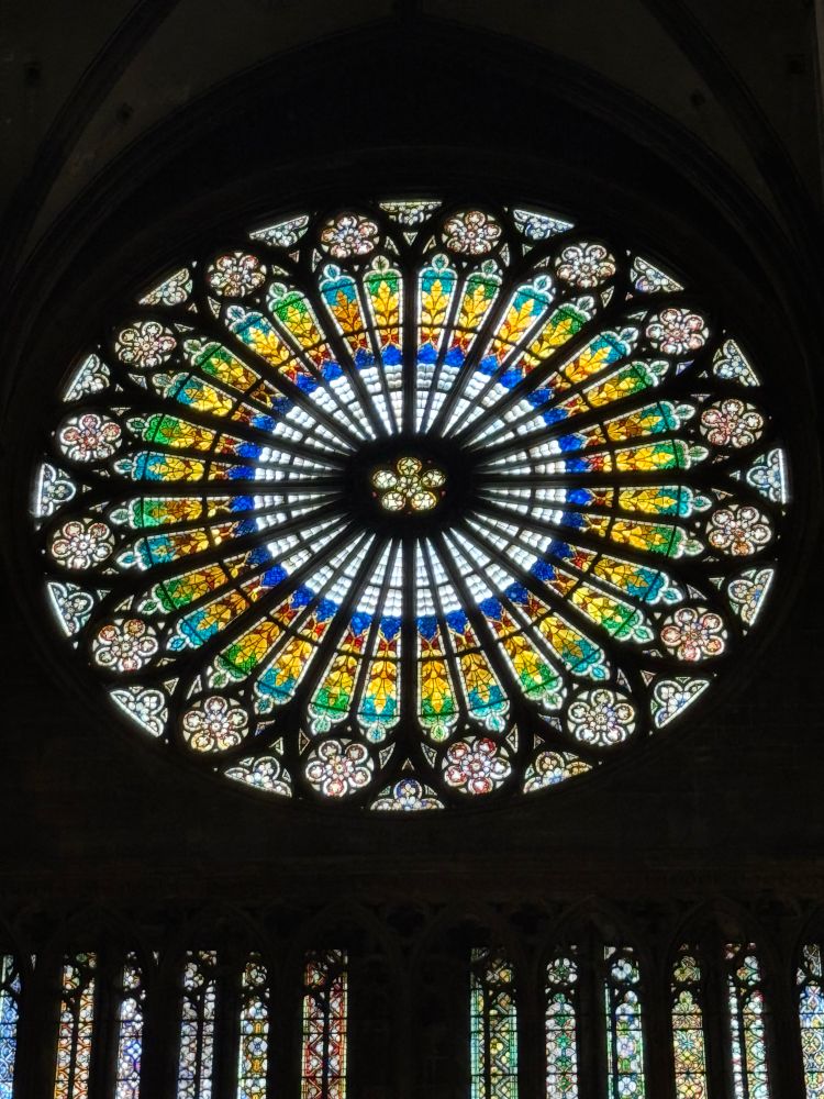 Large, circular, leaded stained glass window with circular designs in the center and around the edges that bring to mind geometric flowers. Concentric circles radiate from the center in black, white, blue. Past the blue circle are panels in yellow, brown, and green that represent crops (it looks like it could be wheat). Below the window are 12 smaller elongated stained glass windows in a variety of colors and designs, mostly evoking flowers. 