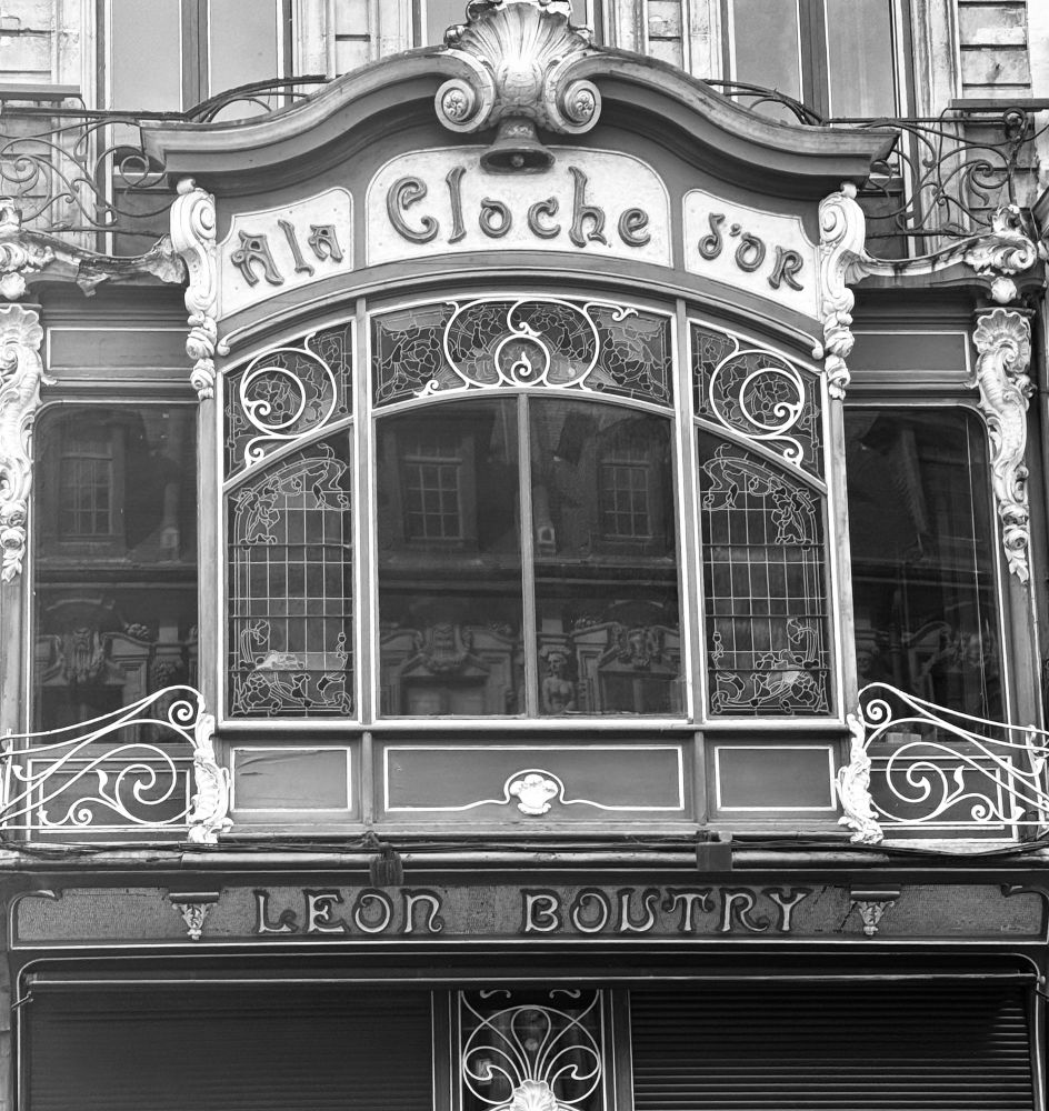 Early 20th century building boasts an elegant window with curly wrought iron embellishments. The name of a shop that is no longer there is at the top: À la Cloche d’or