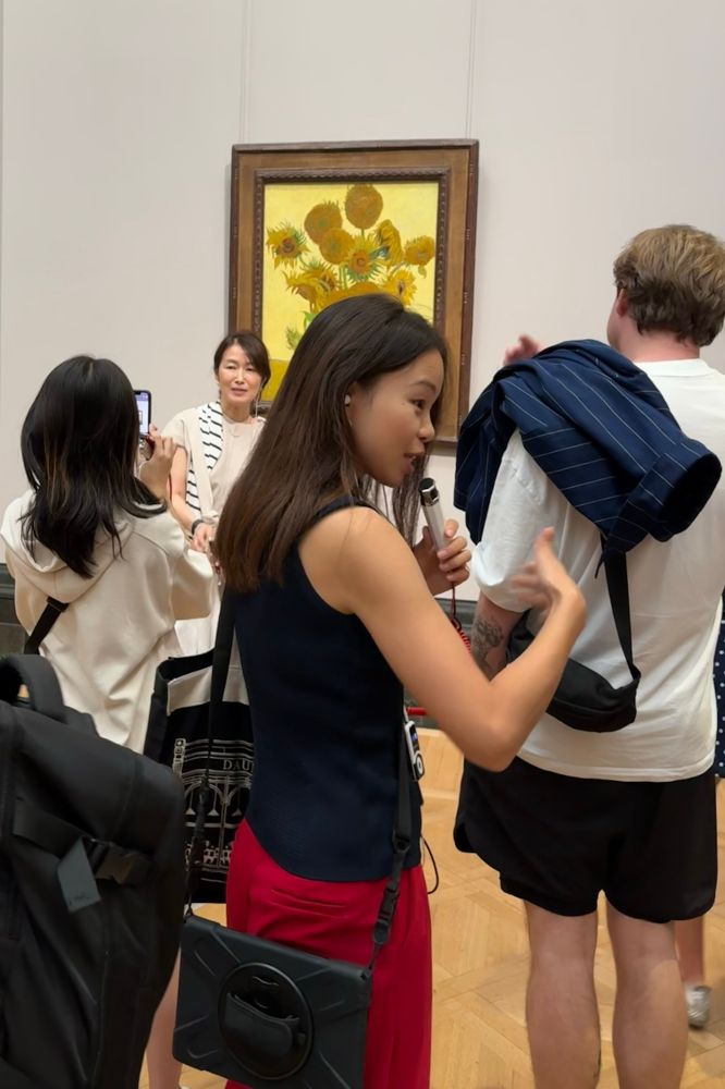 Scene in front of Van Gogh’s Sunflowers painting. Directly in front of the camera is a tour guide speaking into a microphone. She is turned to the side, gesturing as she talks. To the right is a man in a white T and shorts, a “fanny pack” dangling by its strap from his shoulder to rest on his back. A woman stands in front of the painting, smiling, while another woman uses a phone to take her picture. 