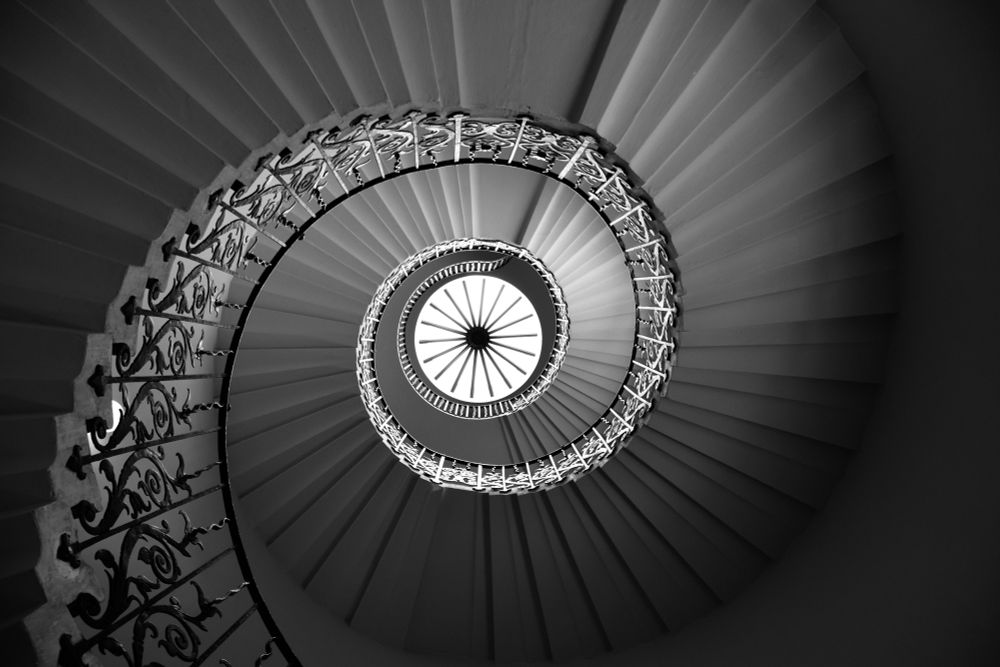 Black and white photo of the Tulip Stairs in the Queen’s House, Greenwich. This view is underneath the circular staircase, looking up. It is dark around the edges with a spiral of light that leads to a window, a circle of light with a dark center and spokes.
