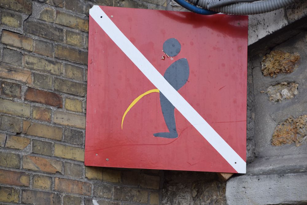A square, red metal sign with a diagonal white stripe across a cartoon silhouette of a man peeing (an arced yellow line emerges from under the white stripe). 