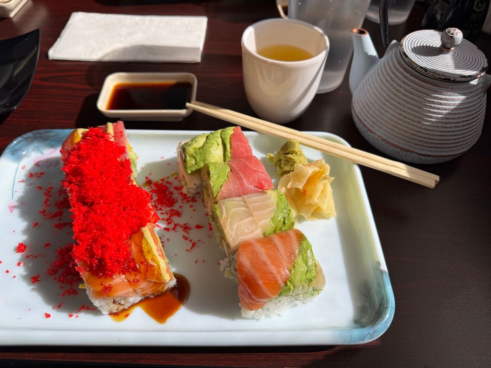 Sunlit sushi rolls on a white rectangular tray with tinges of blue. One roll is topped with red crumbles, with salmon and eel sauce visible. The other is a rainbow roll, with salmon, yellow tail, tuna, and avocado on top. Ginger, wasabi, and bamboo chopsticks are at one of the tray’s corners. Also on the table are a white teapot, a white cup holding green tea, and a small rectangular bowl holding soy sauce. 