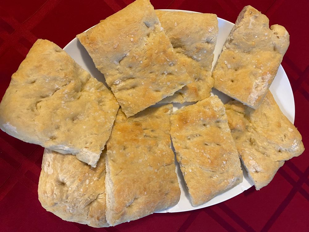 Seen from above. A white oval platter on a red tablecloth. Eight pieces of plain focaccia are arranged on the platter. One can see the dimples in the golden-brown bread and the Maldon salt flakes sprinkled on top.