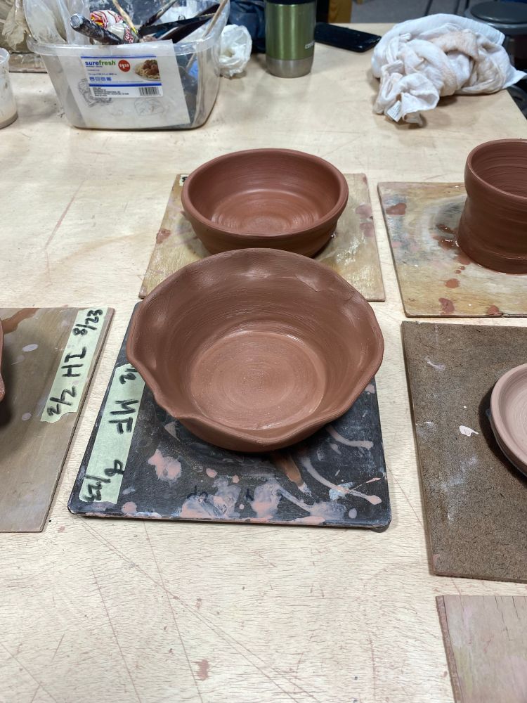 A look at finished but unglazed and unfired pots from class. On a wooden table surface, in the center of the photo are two bowls set on thin wood squares ready to be painted with glaze. One can see the edges of two others along the right side. At the top of the image, from the left, are a plastic tub with tools, a green go-cup, and a white rag.