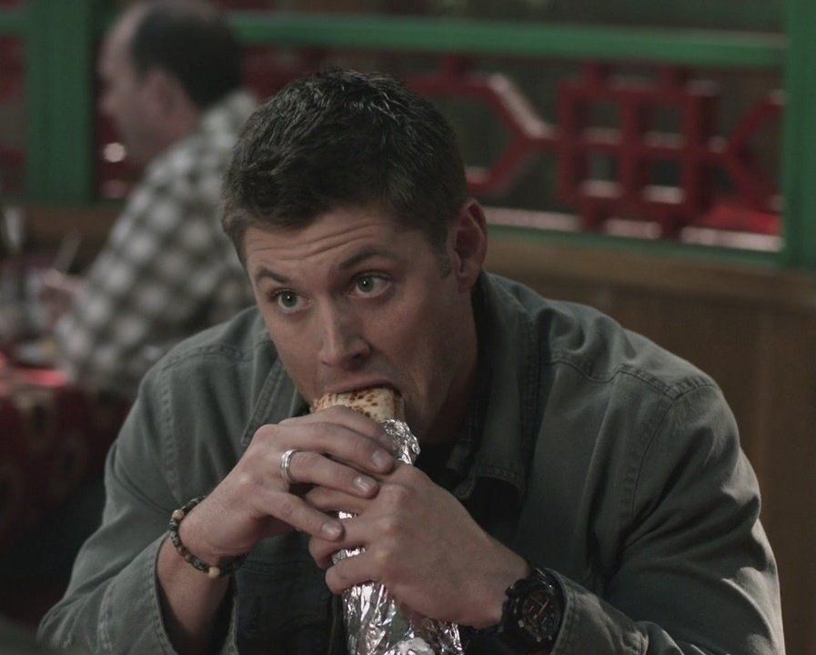 Early seasons Dean with his ring and his bracelet eating a burrito and raising his eyebrows. He's wearing a green outer denim shirt 