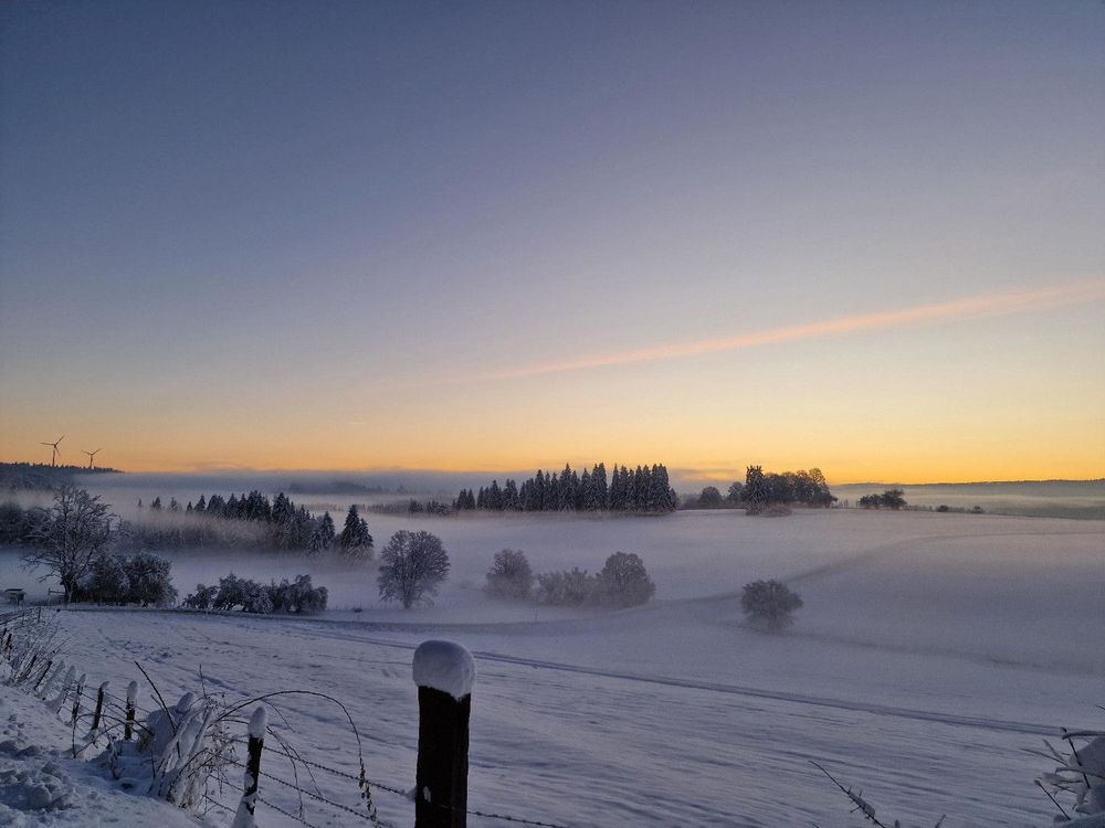Eine Winterlandschaft mit verschneiten Bäumen, Nebel und einem Zaun im Vordergrund, unter einem Himmel mit orangefarbenen Sonnenuntergangfarben.