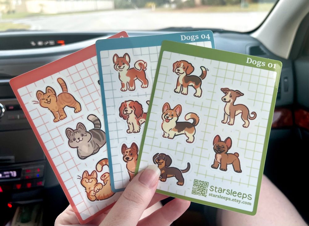 A photo of three sticker sheets. Sheet 1 has small dogs: A Beagle, Corgi, Dachshund, Whippet/Italian Greyhound, and Frenchie. Sheet 2 has medium dogs: a Shiba, Cocker Spaniel, and Blue Heeler. Sheet 3 has cats: two orange tabbies and one grey tabby. 
